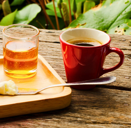 A red mug of coffee and a glass of honey drink sit on a wooden table, with lush green foliage in the background.の写真素材
