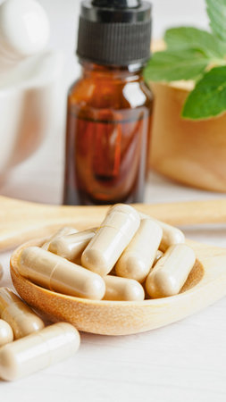 A wooden spoon holds numerous capsules. In the background, a dark glass bottle and fresh green leaves suggest natural remedies.の写真素材