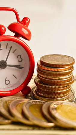A vintage red alarm clock sits beside a pile of gold coins, symbolizing the passage of time and wealth.の写真素材