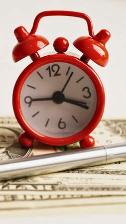 A vintage red alarm clock rests on a stack of bills, with a pen in the foreground, suggesting time and finances.の写真素材