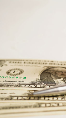 A detailed view of stacked US dollar bills with a sleek silver pen positioned across them,...
