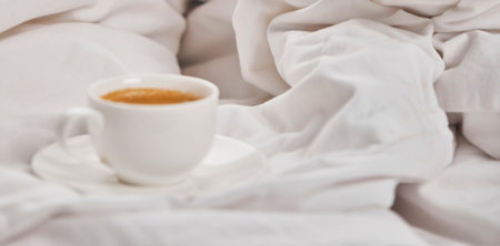 A warm cup of coffee rests on a saucer amidst soft, rumpled white linens, suggesting a peaceful morning.の写真素材