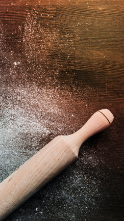 A light-colored wooden rolling pin rests on a dark, textured surface dusted with white flour, suggesting baking preparation.の写真素材