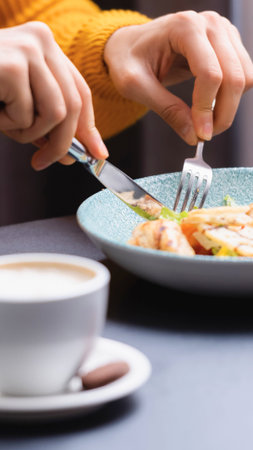 Close up of hands using cutlery to eat a meal from a plate, with a coffee cup in the foreground.