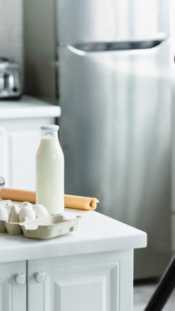 A glass bottle of milk and a carton of eggs sit on a white kitchen counter next to a stainless steel refrigerator.の写真素材