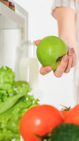 A hand holds a bright green apple above a selection of fresh produce, including lettuce, tomatoes, and cucumber, inside a refrigerator.の写真素材
