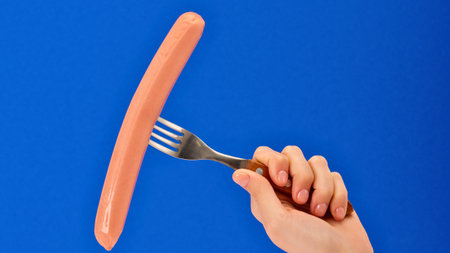 A person's hand grips a fork, piercing a long, uncooked sausage. The background is a solid, vibrant blue.の写真素材
