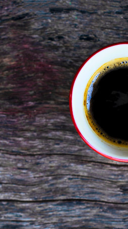 A close-up shot of a steaming cup of dark coffee with a reddish rim, resting on a textured, weathered wooden surface.の写真素材