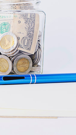 A close-up shot shows a glass jar filled with coins and banknotes, resting on a white surface next...