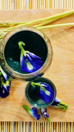 Close-up of vivid blue butterfly pea flowers and buds arranged on a textured wooden surface with dried grass stalks.の写真素材