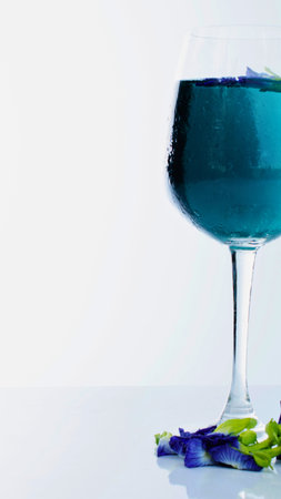 A striking blue beverage, likely made from butterfly pea flowers, is presented in a stemmed glass with scattered petals on a reflective surface.の写真素材