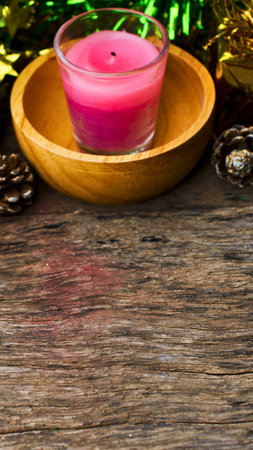A lit pink candle in a glass jar sits on a wooden plate, surrounded by pinecones and festive green and gold tinsel.の写真素材