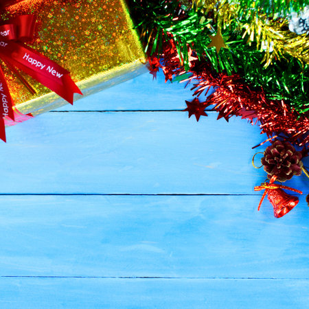 A golden gift box with a red ribbon sits beside colorful Christmas tinsel and garland on a rustic blue wooden surface.の写真素材