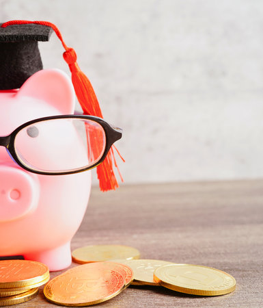 A pink piggy bank dressed as a graduate sits with coins, symbolizing financial education and savings for future success.の写真素材