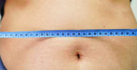 A close-up shot shows a blue measuring tape wrapped around a person's bare midsection, indicating waist size.の写真素材