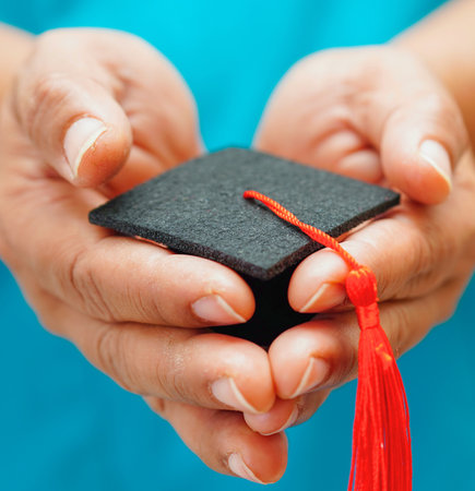 Close-up of two hands gently cradling a small black graduation cap with a vibrant red tassel, symbolizing achievement.の写真素材