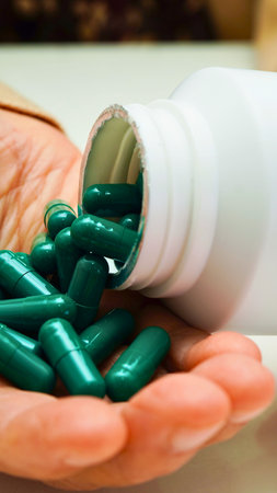 A close-up shot shows a hand holding many shiny green capsules that have spilled from a white prescription bottle.の写真素材