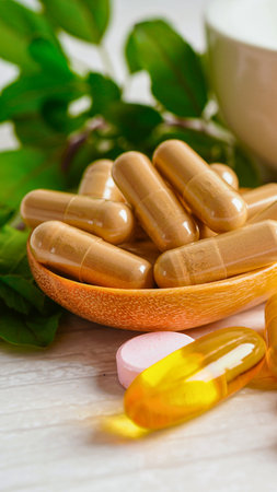 A close-up shot of herbal capsules and oil supplements arranged on a wooden spoon, with fresh green leaves in the background.の写真素材