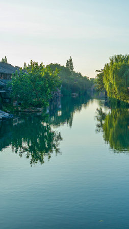 A serene river flows through a landscape of dense green trees, their reflections mirrored perfectly in the calm water under a bright sky.の写真素材
