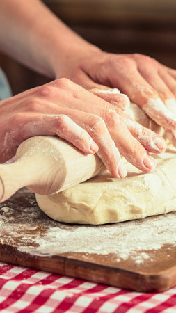 Close-up of hands pressing and shaping raw dough on a rustic wooden surface, with a checkered cloth visible below.の写真素材