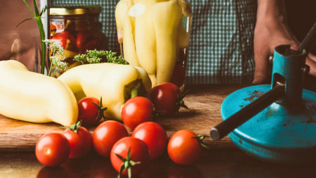 A collection of ripe tomatoes, bell peppers, and jars of preserves on a wooden surface, suggesting homemade food preparation.の写真素材