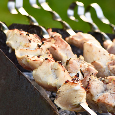 Close-up of seasoned chicken pieces skewered and cooking on a grill, with a blurred green background.の写真素材