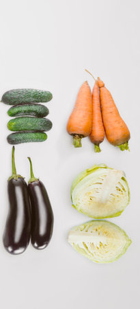 A collection of raw vegetables including dark purple eggplants, orange carrots, green cucumbers, and a head of cabbage, presented on a white background.の写真素材