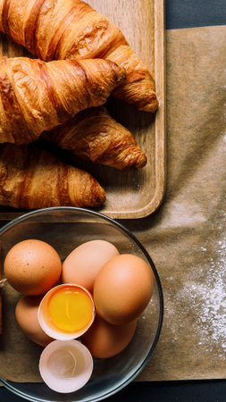 A close-up view of a stack of golden brown croissants and a bowl of fresh eggs, one cracked open revealing its yolk.の写真素材
