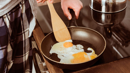 A person is cooking an egg in a black frying pan with a wooden spatula, preparing breakfast in the kitchen.の写真素材