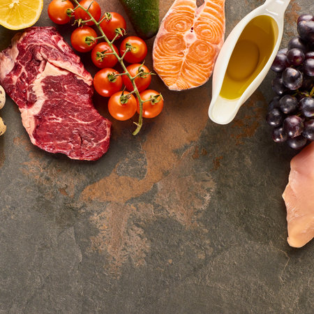 A top-down view showcases a variety of raw ingredients including steak salmon chicken grapes tomatoes and lemon on a textured dark background.の写真素材