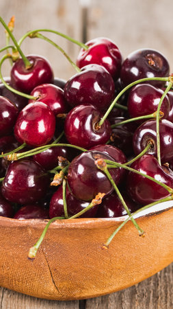 A close-up shot of a pile of glistening, dark red cherries with green stems, nestled in a rustic wooden bowl.の写真素材