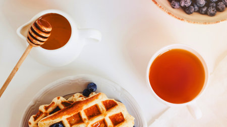 A close-up overhead view of a golden waffle topped with blueberries, served with a honey dipper and a cup of amber liquid.の写真素材