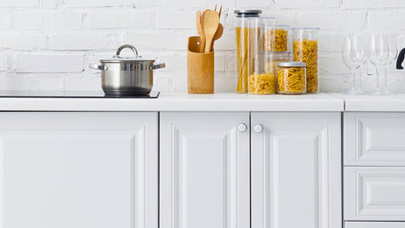 A clean kitchen counter displays glass jars filled with pasta and spices, alongside wooden cooking utensils and a pot.の写真素材
