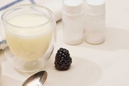 A clear glass of creamy yogurt drink sits next to a single blackberry and a spoon, with two small bottles in the background.の写真素材