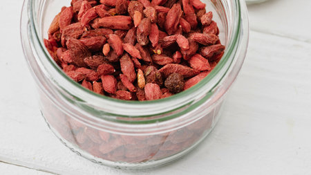 A clear glass jar filled with dried red goji berries sits on a white wooden surface, showing texture and detail.の写真素材