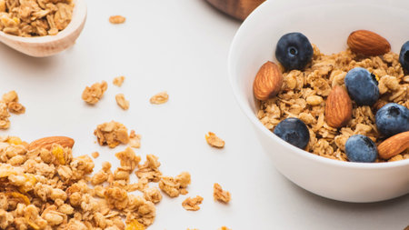 A close-up shot of a white bowl filled with crunchy granola, topped with fresh blueberries and...