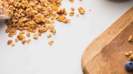 A pile of crunchy granola and a few blueberries are scattered on a white surface next to a wooden cutting board.の写真素材