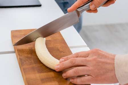 A close-up shot shows hands holding a knife and slicing a banana on a wooden cutting board.の写真素材