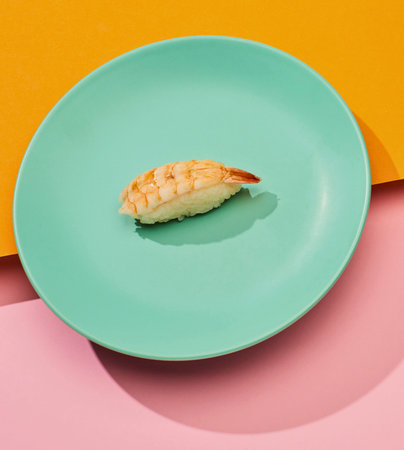 A single piece of cooked shrimp sushi rests on a teal plate against a vibrant pink and orange background.の写真素材