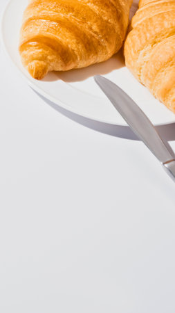 Two golden brown croissants sit on a white surface, with a pair of silver cutlery resting beside them, suggesting a delicious breakfast.の写真素材