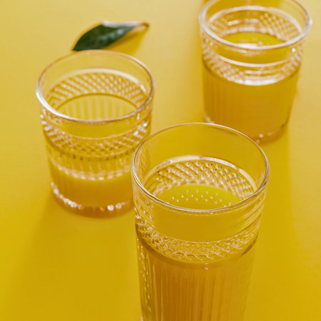 Three glasses filled with vibrant yellow citrus beverages, possibly juice or a cocktail, are arranged on a solid yellow background with a single green leaf.の写真素材