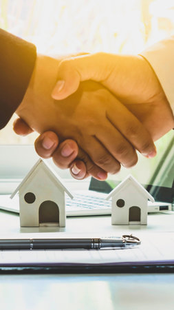 A handshake signifies a successful real estate transaction, with miniature houses and a pen on a desk.の写真素材