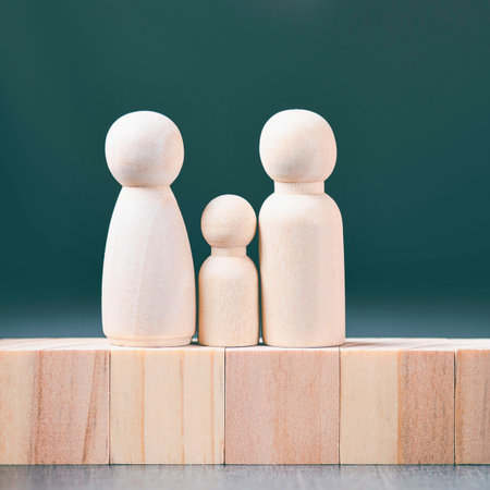 Three simple wooden peg dolls, two larger and one smaller, stand on a row of wooden building blocks against a dark green background.の写真素材