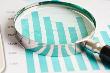 A magnifying glass is placed over a teal bar chart, highlighting the analysis of financial data and trends.の写真素材