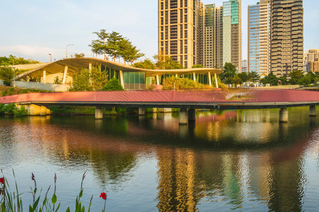 A contemporary red pedestrian bridge spans a calm body of water, reflecting the modern city skyline and lush greenery under a clear sky.の写真素材