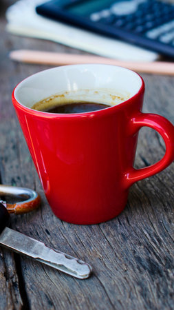A vibrant red coffee cup sits on a weathered wooden surface, holding a steaming cup of dark coffee, with a spoon and keys nearby.の写真素材