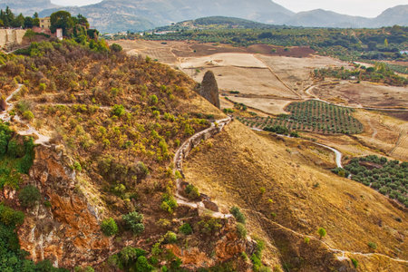 A historic town perched precariously on a steep cliff, overlooking a vast, dry landscape with winding paths and sparse vegetation.の写真素材