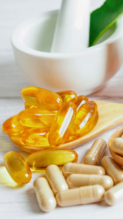 A close-up shot of translucent golden fish oil capsules and opaque white pills scattered on a wooden surface, with a mortar and pestle in the background.の写真素材