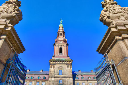A grand, ornate tower with a spire rises majestically between two stone pillars and ornate gates under a vibrant blue sky.の写真素材