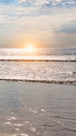 A tranquil beach scene at sunset with the sun casting a golden glow on the ocean waves and wet sand.の写真素材